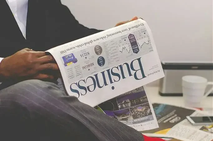 Mann in Anzug liest eine Zeitung, neben ihm stehen Pflanzen und ein Kaffeebecher.