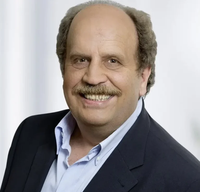 Lächelnder Mann mit dunkler Blazer und hellblauer Hemd, vor neutralem Hintergrund.