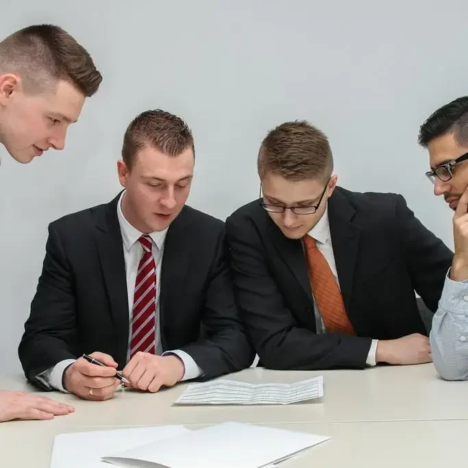Vier Männer in Anzügen arbeiten konzentriert an einem Tisch und betrachten Dokumente.
