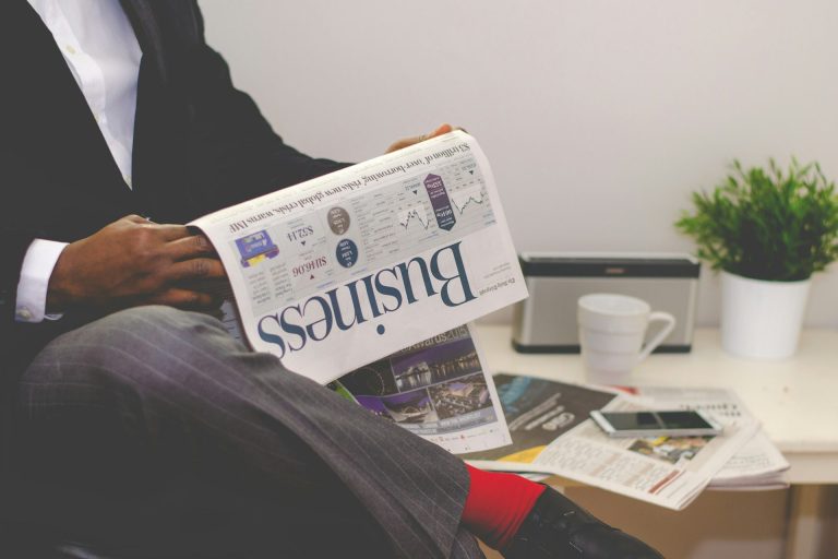 Mann in Anzug liest eine Zeitung, neben ihm stehen Pflanzen und ein Kaffeebecher.