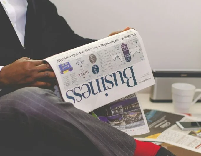 Mann in Anzug liest eine Zeitung, neben ihm stehen Pflanzen und ein Kaffeebecher.