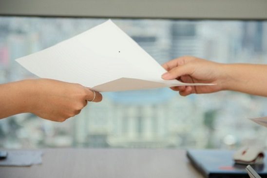Zwei Hände übergeben ein Blatt Papier vor einem Fenster mit Stadtblick.