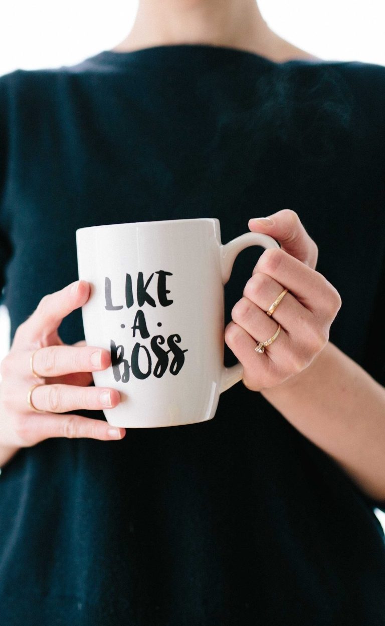 Beige Tasse mit dem Aufdruck "LIKE A BOSS", gehalten von einer Person mit schwarzem Oberteil.