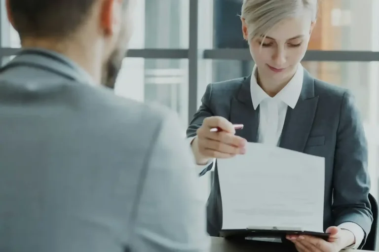 Frau in Anzug reicht Dokument an einen Mann in grauem Anzug.
