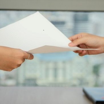 Zwei Hände reichen sich ein Blatt Papier über einen Tisch mit Stadtansicht im Hintergrund.